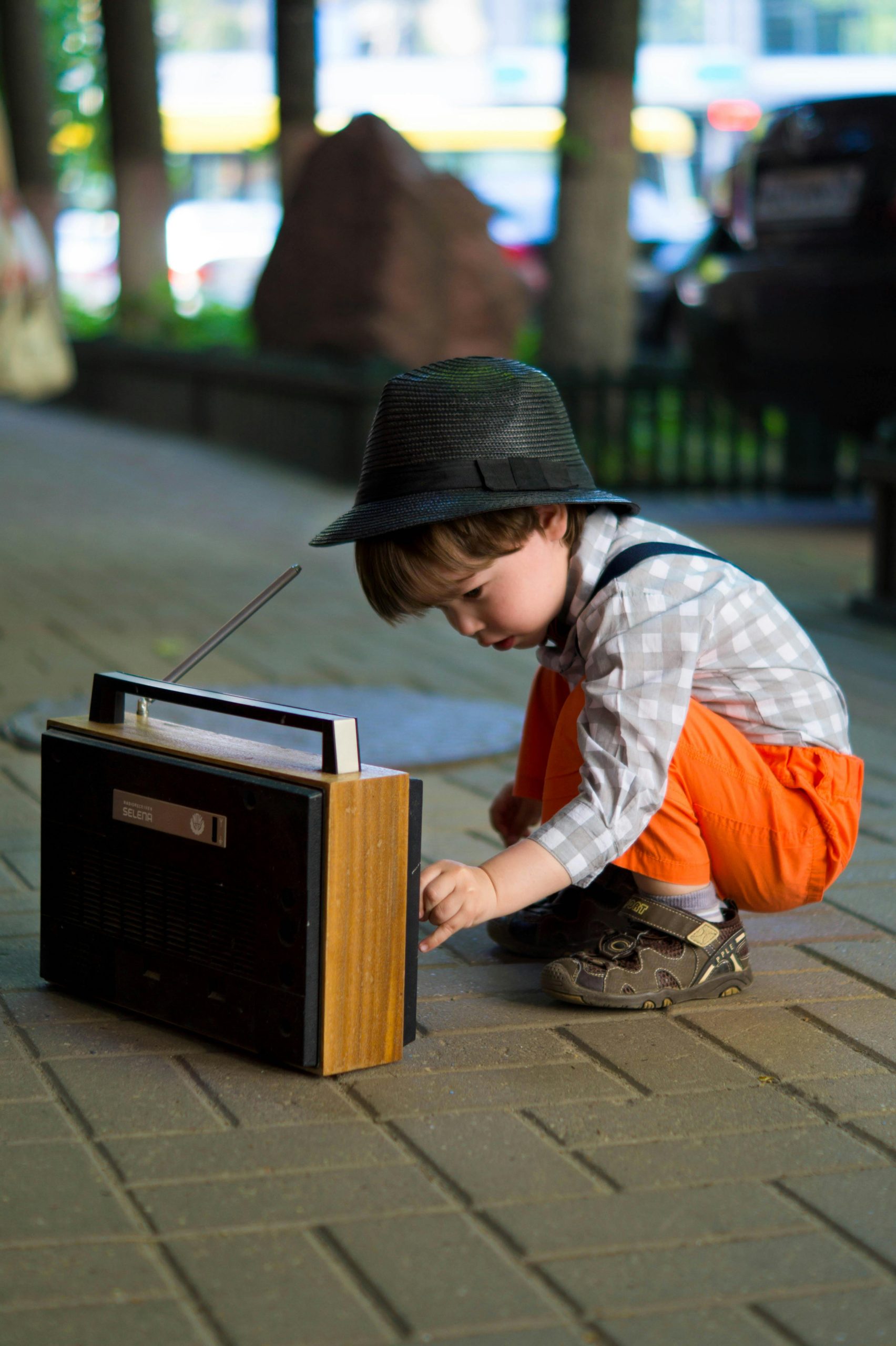 A young boy in fashionable attire adjusts an old-fashioned radio outdoors.